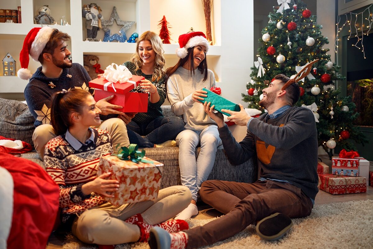Mai mulți prieteni stau lângă bradul de Crăciun și își oferă cadouri în timp ce sunt îmbrăcați cu căciuli de elfi și coarne de ren