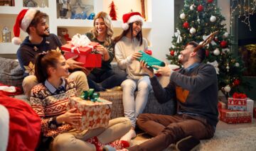 Mai mulți prieteni stau lângă bradul de Crăciun și își oferă cadouri în timp ce sunt îmbrăcați cu căciuli de elfi și coarne de ren