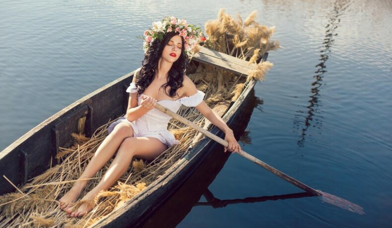 Fată frumoasă stă într-o barcă cu o vâslă și o coroană de flori