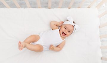 O fetiță frumoasă într-un body alb în timp ce zâmbește și râde din pătuțul său