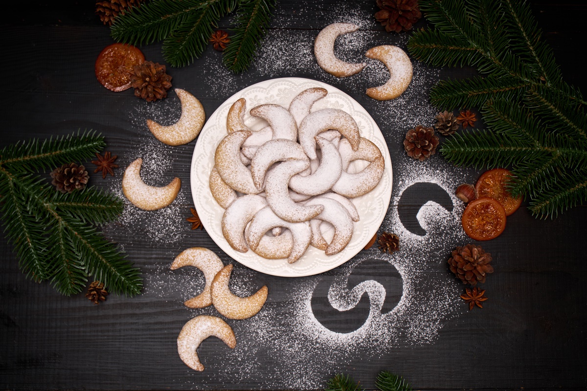 Cornulețe cu nucă și vanilie pe un platou rotund, alături de crengi de brad, conuri, batoane de scorțișoară și anason stelat