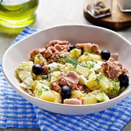 Un bol alb de salată cu ton și cartofi fierți, măsline, perfecte pentru a ști cum să mănânci mai sănătos în anul 2023, așezat pe un prosop în carouri alb-albastre