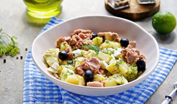 Un bol alb de salată cu ton și cartofi fierți, măsline, perfecte pentru a ști cum să mănânci mai sănătos în anul 2023, așezat pe un prosop în carouri alb-albastre
