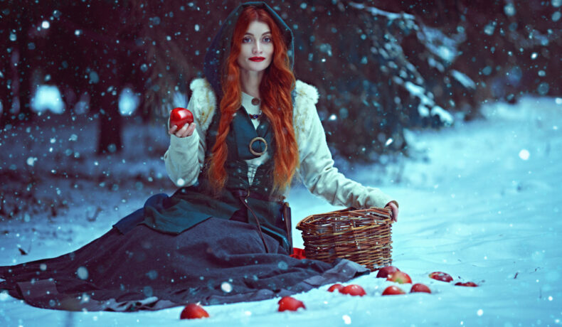 Fată frumoasă cu părul lung stă așezată în zăpadă cu un măr în mână