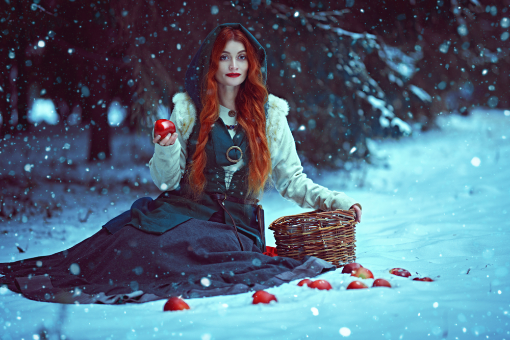 Fată frumoasă cu părul lung stă așezată în zăpadă cu un măr în mână