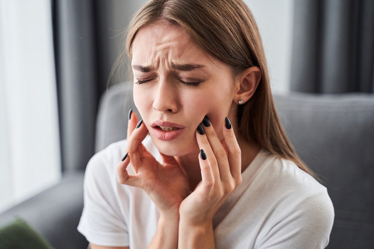 O femeie caucaziană stă încruntată cu mâinile pe obraji în timp ce se plânge de dureri insuportabile de dinți
