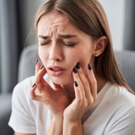 O femeie caucaziană stă încruntată cu mâinile pe obraji în timp ce se plânge de dureri insuportabile de dinți