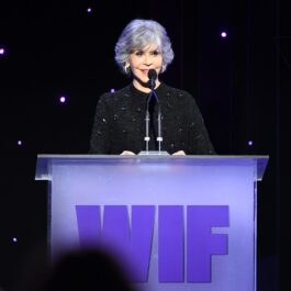 Jane Fonda ține un discurs în spatele unui pupitru de culoare mov, iar actrița este îmbrăcată într-o ținută neagră