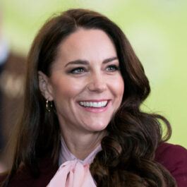 Kate Middleton poartă o pereche de cercei din aur, un sacou vișiniu și o bluză din mătase de culoare roz pal cu un detaliu dintr-o fundă în jurul gâtului.