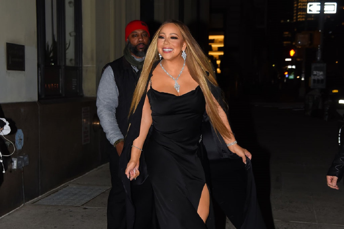 Artista Mariah Carey zâmbește în fața camerelor în timp ce poartă o rochie neagră cu bretele și pestea aceasta o pelerină neagră