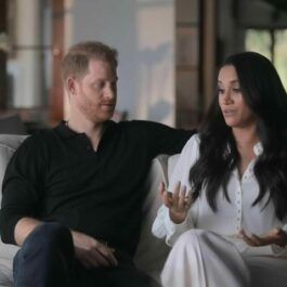 Meghan stă alături de soțul ei pe o canapea albă în timp ce gesticulează în cadrul unui interviu