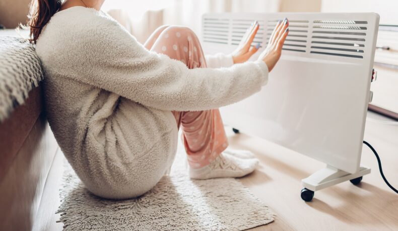 O femeie îmbrăcată gros în pijamale care încearcă să-și încălzească mâinile și picioarele