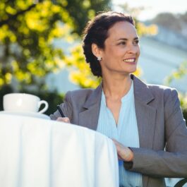 O femeie de afaceri sofisticată, cu părul prins în coadă poartă haine stil business și lucrează pe o terasă, în fața unei cești de cafea de culoare albă