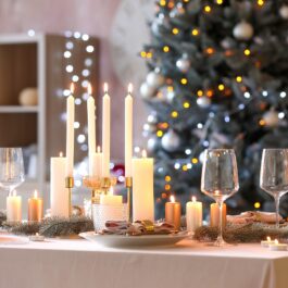 O masă de culoare albă are deaupra ei mai multe pahare, platouri și lumânări pentru o cină de Crăciun, iar pe fundal se află un brad împodobit și o bibliotecă