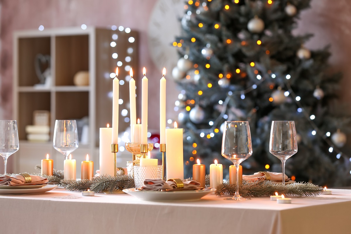 O masă de culoare albă are deaupra ei mai multe pahare, platouri și lumânări pentru o cină de Crăciun, iar pe fundal se află un brad împodobit și o bibliotecă