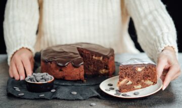 Prăjitură cu mere și ciocolată porționată pe o farfurie, alături de platoul cu prăjitură și un bol cu bănuți de ciocolată