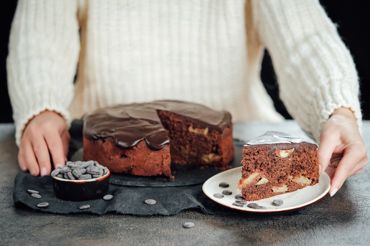Prăjitură cu mere și ciocolată porționată pe o farfurie, alături de platoul cu prăjitură și un bol cu bănuți de ciocolată