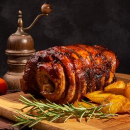 Friptură din fleică de porc rulată, pe un suport de lemn, alături de cartofi, crenguțe de rozmarin și o râșniță vitange