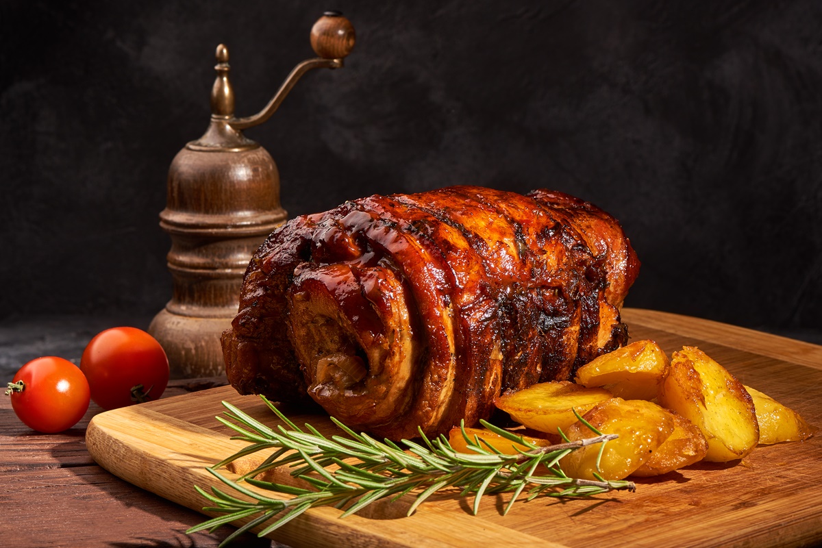 Friptură din fleică de porc rulată, pe un suport de lemn, alături de cartofi, crenguțe de rozmarin și o râșniță vitange