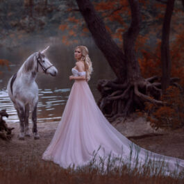 Fată frumoasă îmbrăcată într-o rochie roz lungă stă într-o pădure lângă un cal