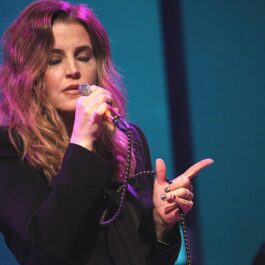 Lisa Marie Presley poartă o bluză de culoare neagră, părul despletit și ține un microfon în mâna dreaptă în timp ce cântă o melodie