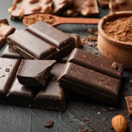 Mai multe bucăți de ciocolată neagră așezate pe un blat gri, alături de un bol de cacao sugerează unul dintre alimentele cu efect antiinflamator