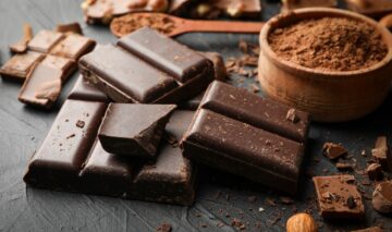 Mai multe bucăți de ciocolată neagră așezate pe un blat gri, alături de un bol de cacao sugerează unul dintre alimentele cu efect antiinflamator