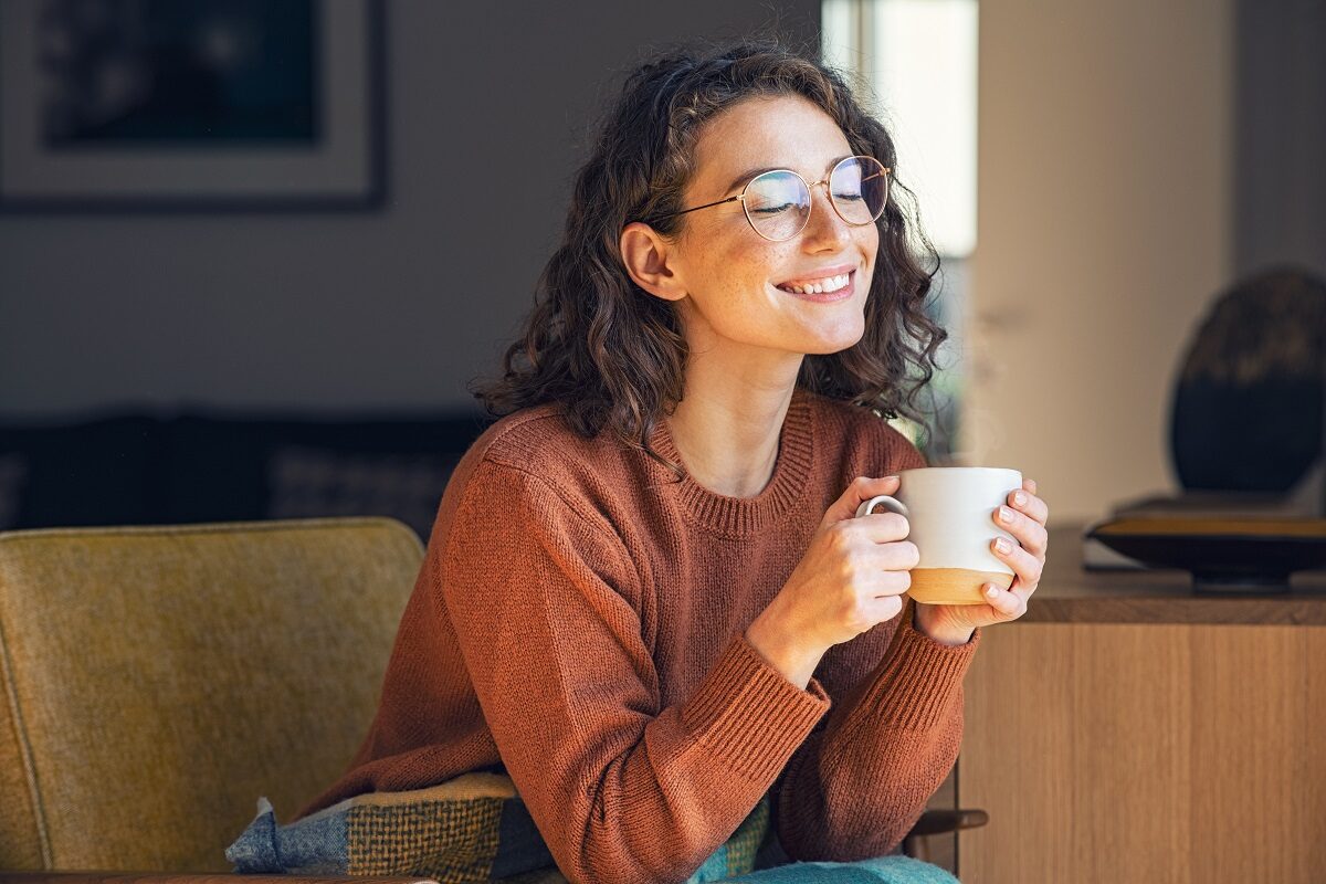 O femeie frumoasă cu părul creț și ochelari care savurează o cană de ceai, fiind pe lista de băuturi care te ajută să ai un abdomen plat