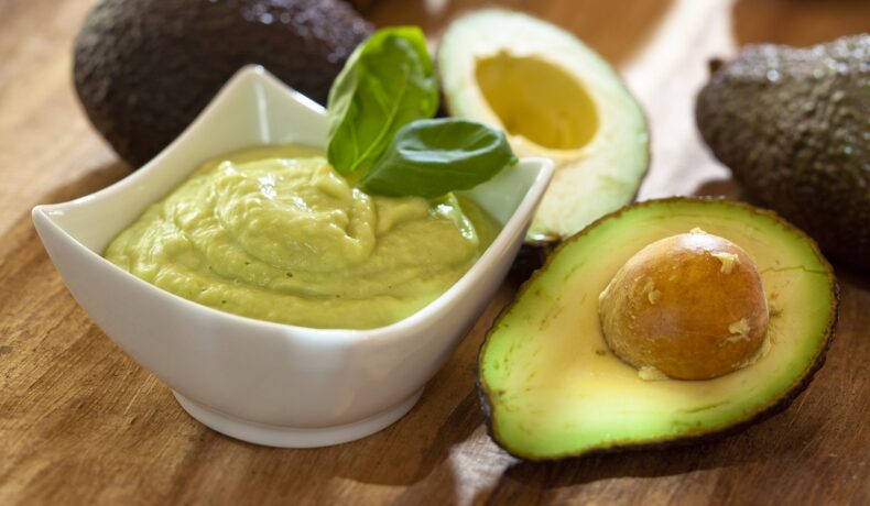 Un avocado tăiat în două, lângă un bol cu pastă de avocado sugerează beneficiile consumului acestui fruct
