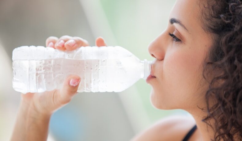 O femeie tânără bea apă dintr-o sticlă de plastic transparentă