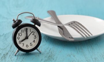 Un ceas desteptător de culoare neagră, alături de o farfurie albă, goală sugerează ce se întâmplă în organism dacă sari peste mese