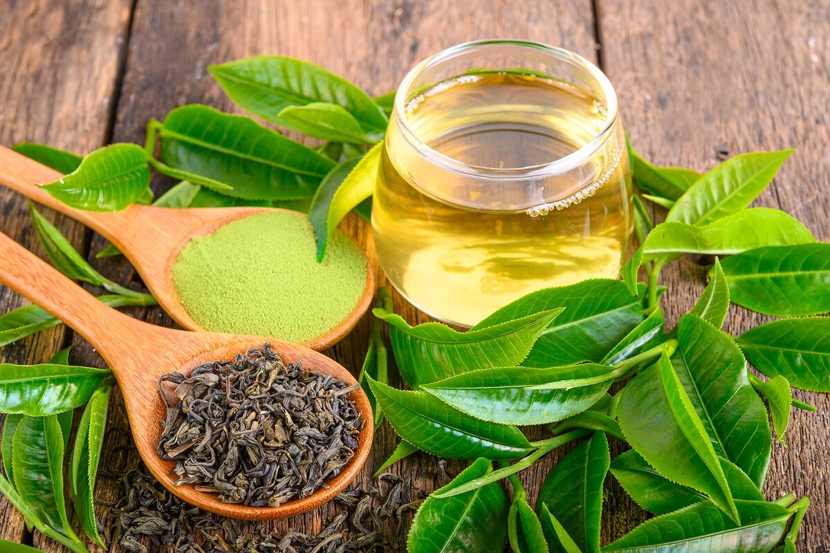 Mai multe frunze de ceai verde, alături de o cană de ceai și o lingură de lemn sugerează unul dintre ceaiurile cu un puternic efect antiinflamator