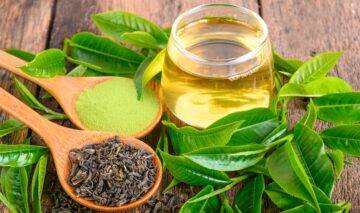 Mai multe frunze de ceai verde, alături de o cană de ceai și o lingură de lemn sugerează unul dintre ceaiurile cu un puternic efect antiinflamator