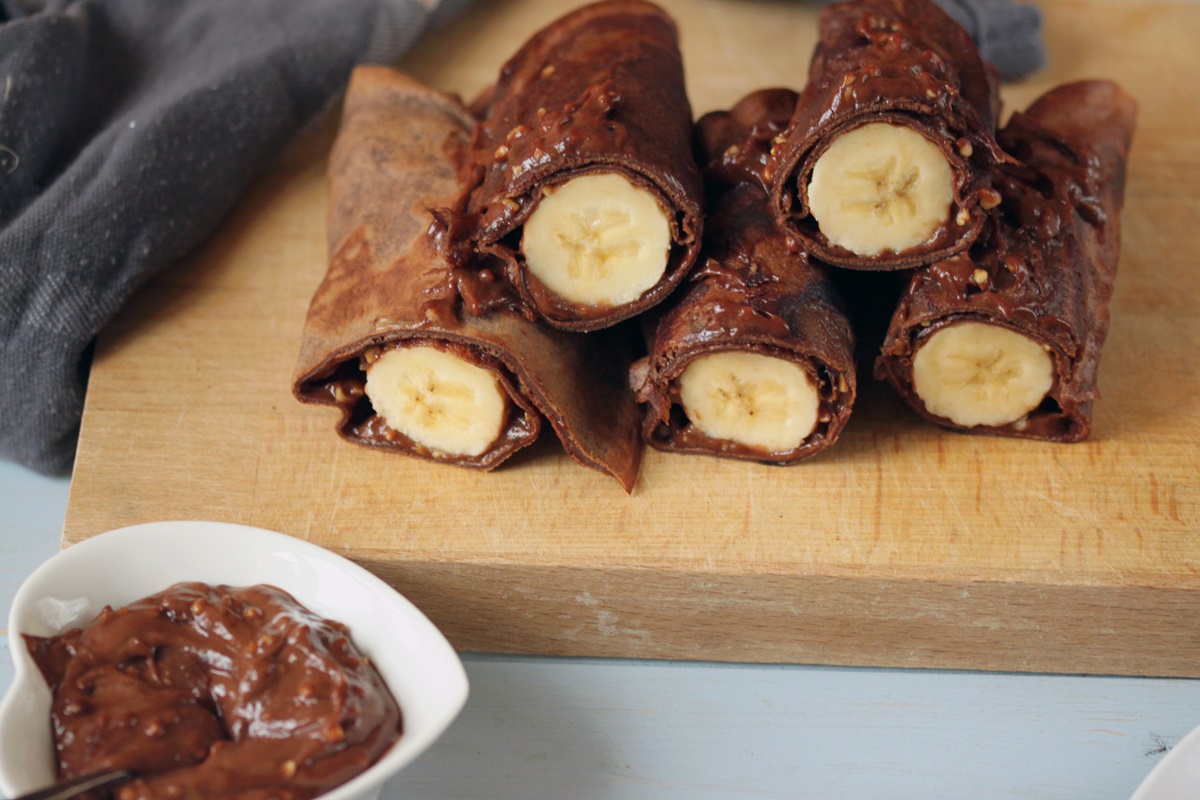 Clătite din aluat cu cacao umplute cu cremă de ciocolată și banane pe un tocător de lemn