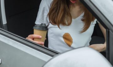 O femeie tânără care se află într-o mașină poartă un tricou alb peste care a scăpat cafea și s-a fosrmat o pastă maro, mare
