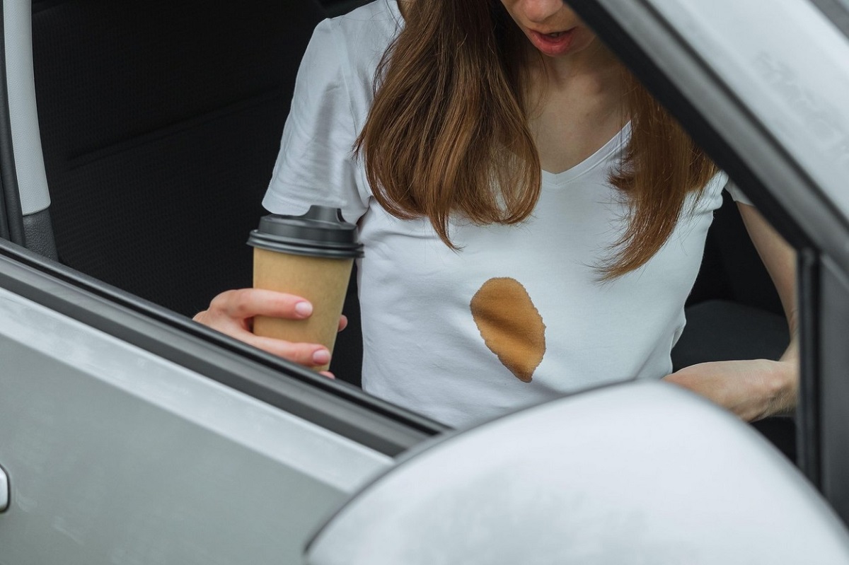 O femeie tânără care se află într-o mașină poartă un tricou alb peste care a scăpat cafea și s-a fosrmat o pastă maro, mare