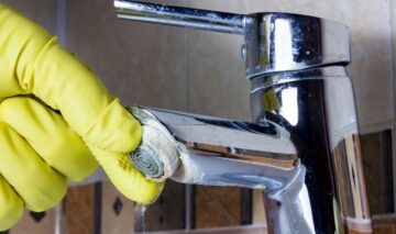 Un robinet de bucătărie plin de calcar și o mână care poartă o mănușă de plastic de culoare galbenă ce urmează să curețe de calcar zona respectivă