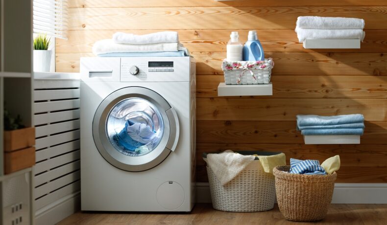 O mașină de spălat de culoare albă este așezată în colțul unei băi și este programată la un program de spălare a rufelor