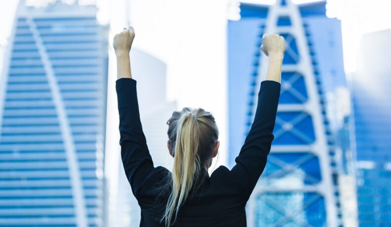O femeie tânără de afaceri se bucură cu mâinile în aer în fața unir clădiri mari de birouri, purtând un de tip business