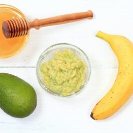 O banană, un avocado, un bol cu avocado amestecat cu banană și un bol cu miere și ustensilă de lemn