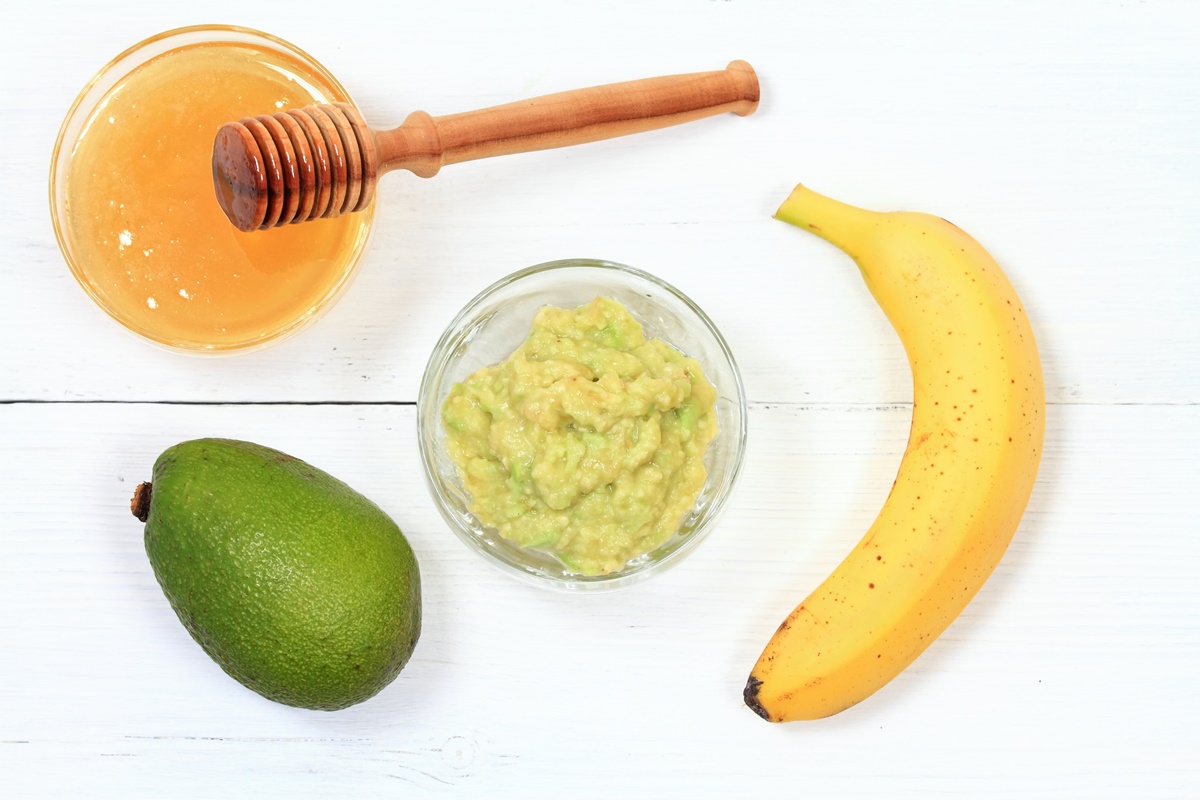 O banană, un avocado, un bol cu avocado amestecat cu banană și un bol cu miere și ustensilă de lemn