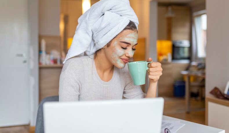 O femeie stă în fața unui laptop, fericită, cu o mască facială și un prosop în cap, sugerând obiceiuri care îți fac viața mai bună