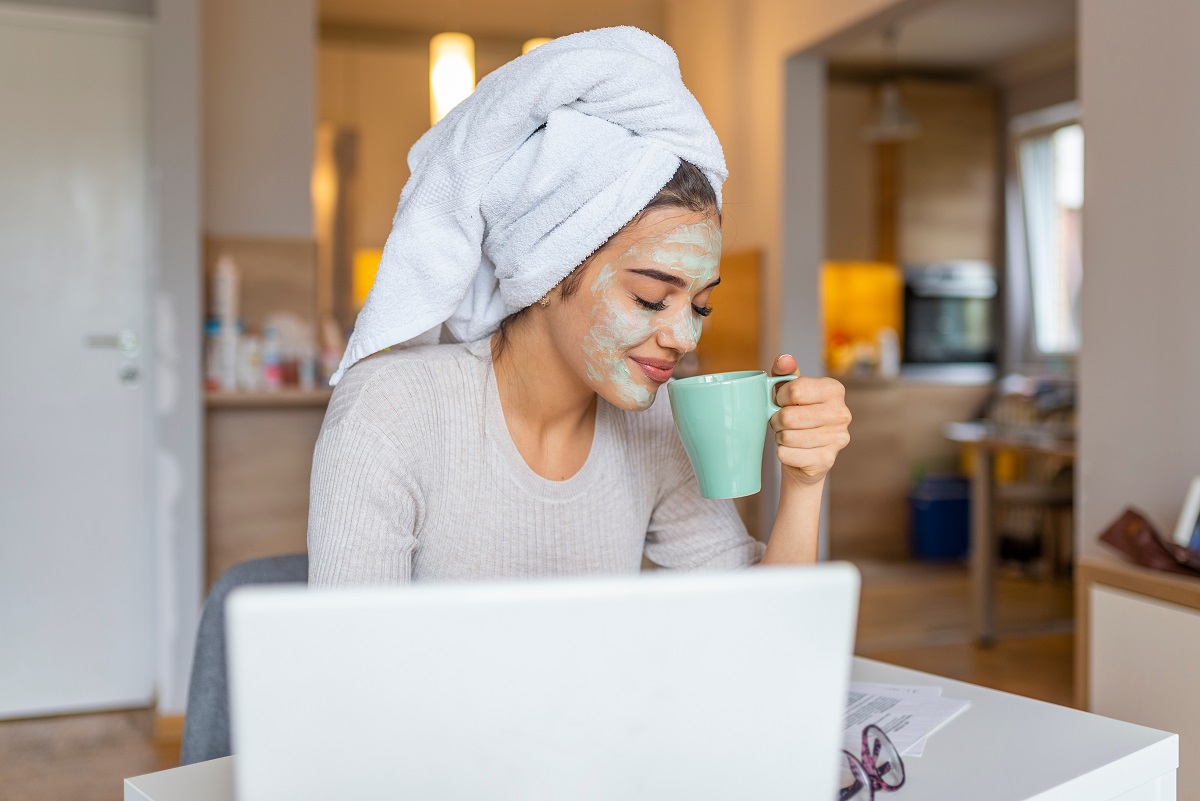 O femeie stă în fața unui laptop, fericită, cu o mască facială și un prosop în cap, sugerând obiceiuri care îți fac viața mai bună