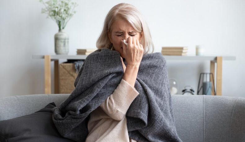 O femeie cu vârsta în jurul a 50 de ani este răcită și stă pe o canapea, cu o pătură pe ea fiindcă nu a respectat obiceiuri care protejează imunitatea în sezonul rece
