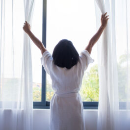 O femeie cu părul negru până la umeri, poartă o pijama și lasă lumina soarelui să pătrundă în casă, activitate care face parte din lista cu obiceiuri de urmat dimineața pentru a fi în formă