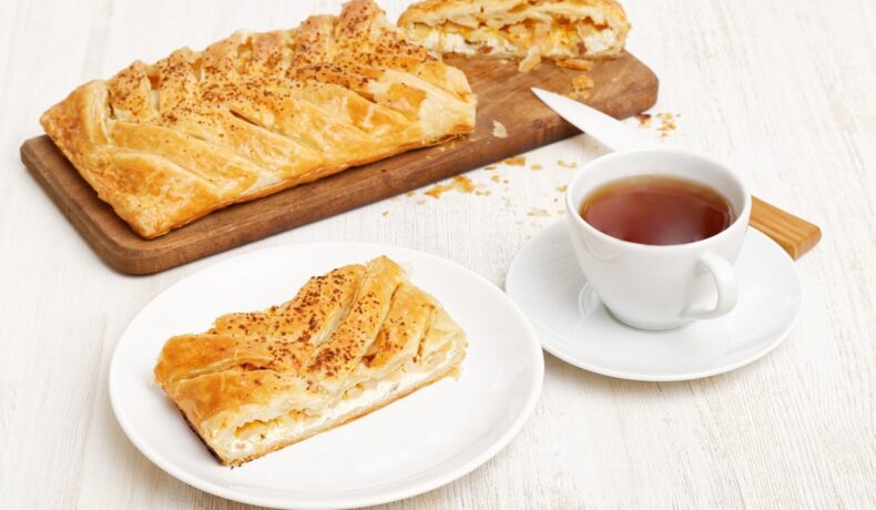 Plăcinte împletite cu brânză și fructe confiate, una porționată pe un tocător de lemn și o porție în farfurie, alături de o ceașcă de ceai