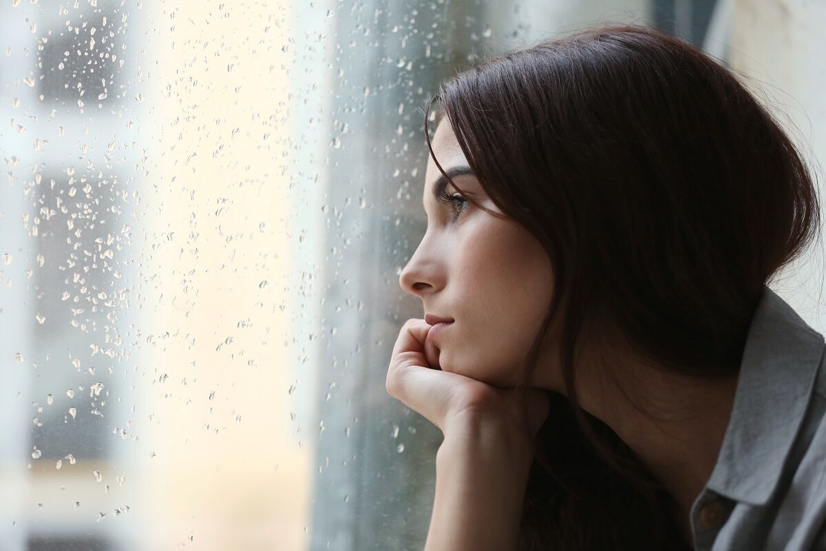 O femeie brunetă și apatică stă în fața unui geam cu mâna în barbă sugerând unul dintre semnele care arată că ești deprimată