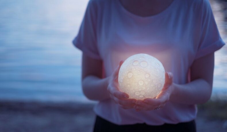 Fată îmbrăcată într-un tricou stă pe plajă și ține în mână o lună mică