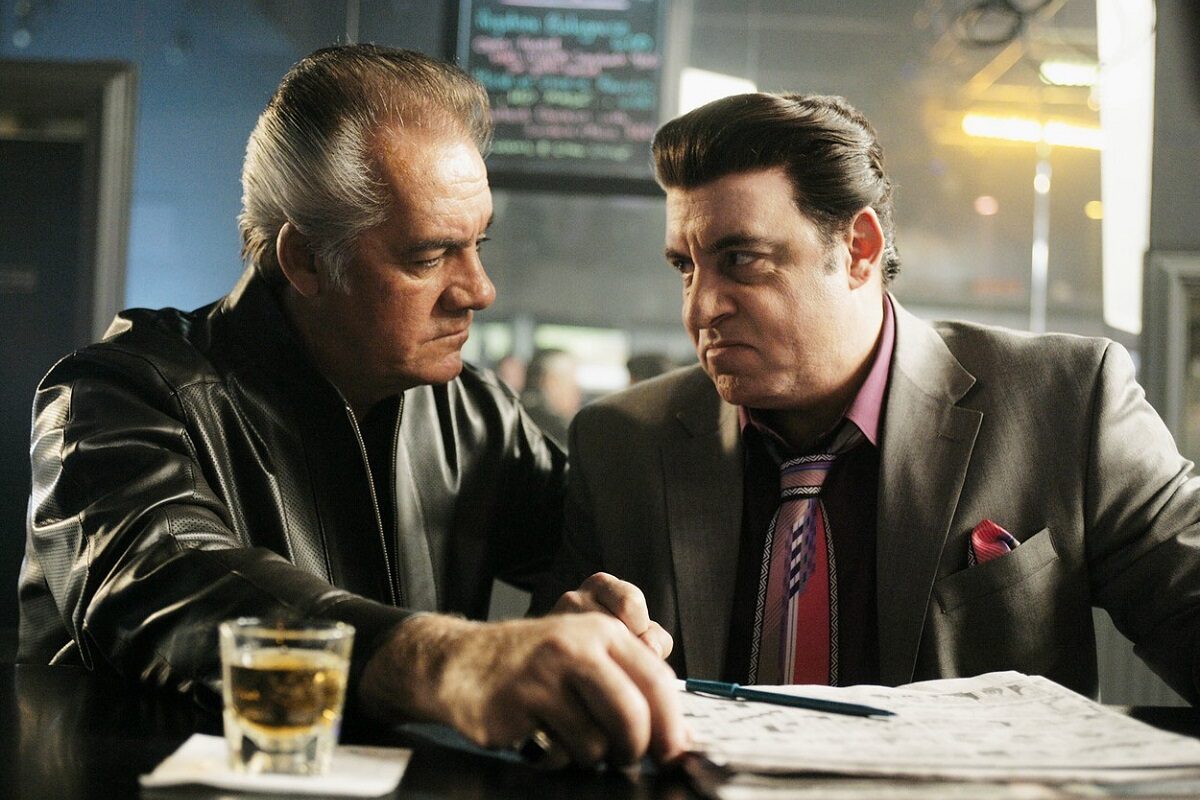 Imagine din serialul „The Sopranos” în care apar James Gandolfini și Tony Sirico îmbrăcați în sacoruri scumpe, stând la o masă în interiorul unui bar