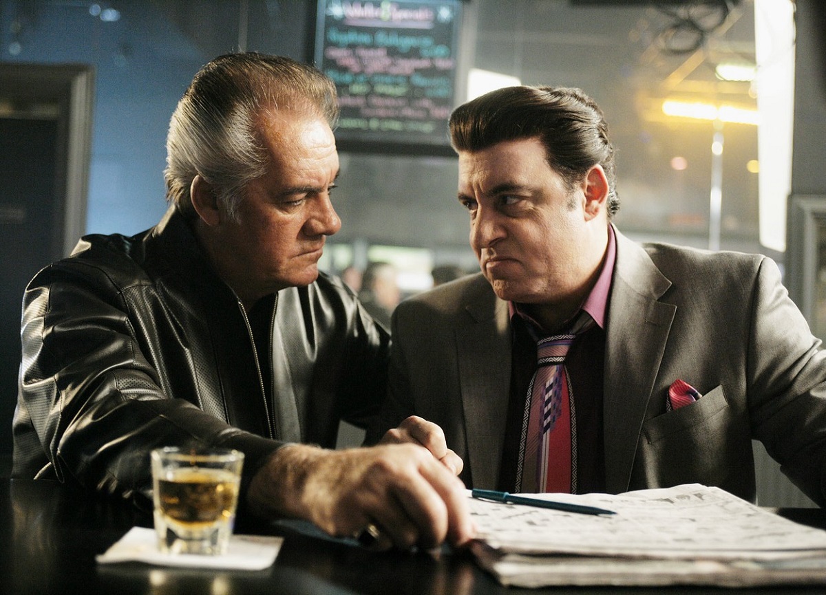 Imagine din serialul „The Sopranos” în care apar James Gandolfini și Tony Sirico îmbrăcați în sacoruri scumpe, stând la o masă în interiorul unui bar